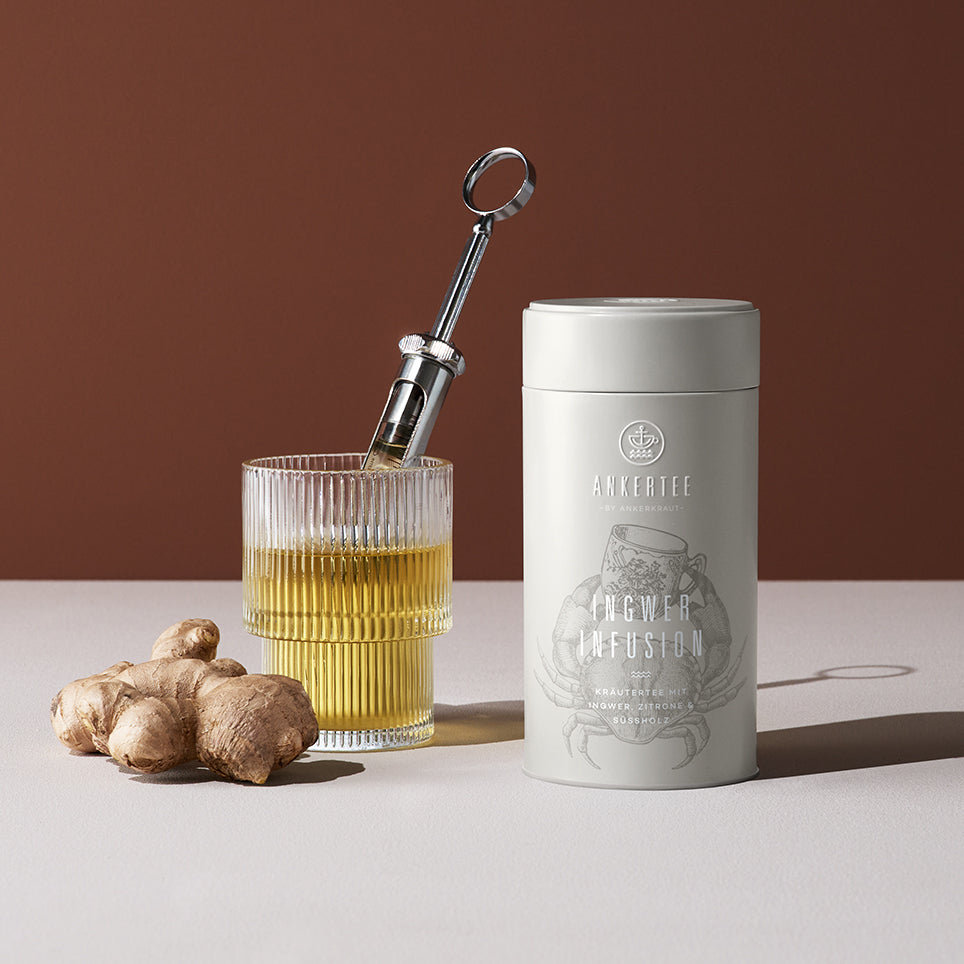 Eine Ankertee-Dose mit der Aufschrift INGWER INFUSION, daneben ein gefülltes Teeglas mit einer Infusionsspritze und davor eine Ingwerknolle