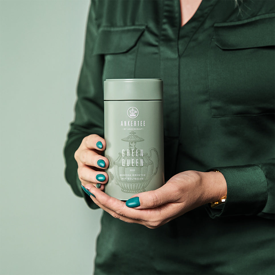 Eine Frau mit grüner Bluse hält in ihren Händen eine Ankertee-Dose mit der Aufschrift GREEN QUEEN.