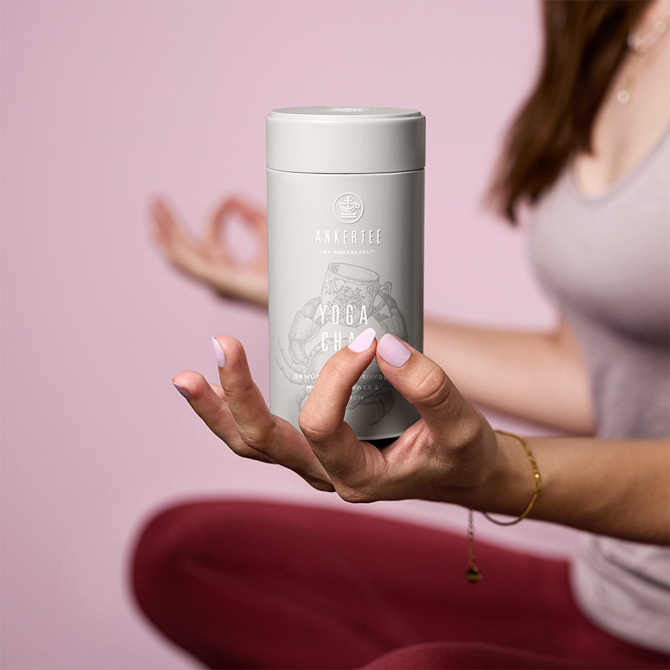 Eine Frau in Yoga-Sitzhaltung hält eine Ankertee-Dose mit der Aufschrift YOGA CHAI