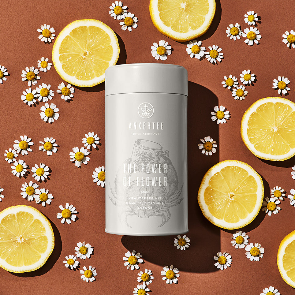 Eine Ankertee-Dose mit der Aufschrift THE POWER OF FLOWER liegt auf einer braunen Fläche umgeben von Blüten und Zitronenscheiben