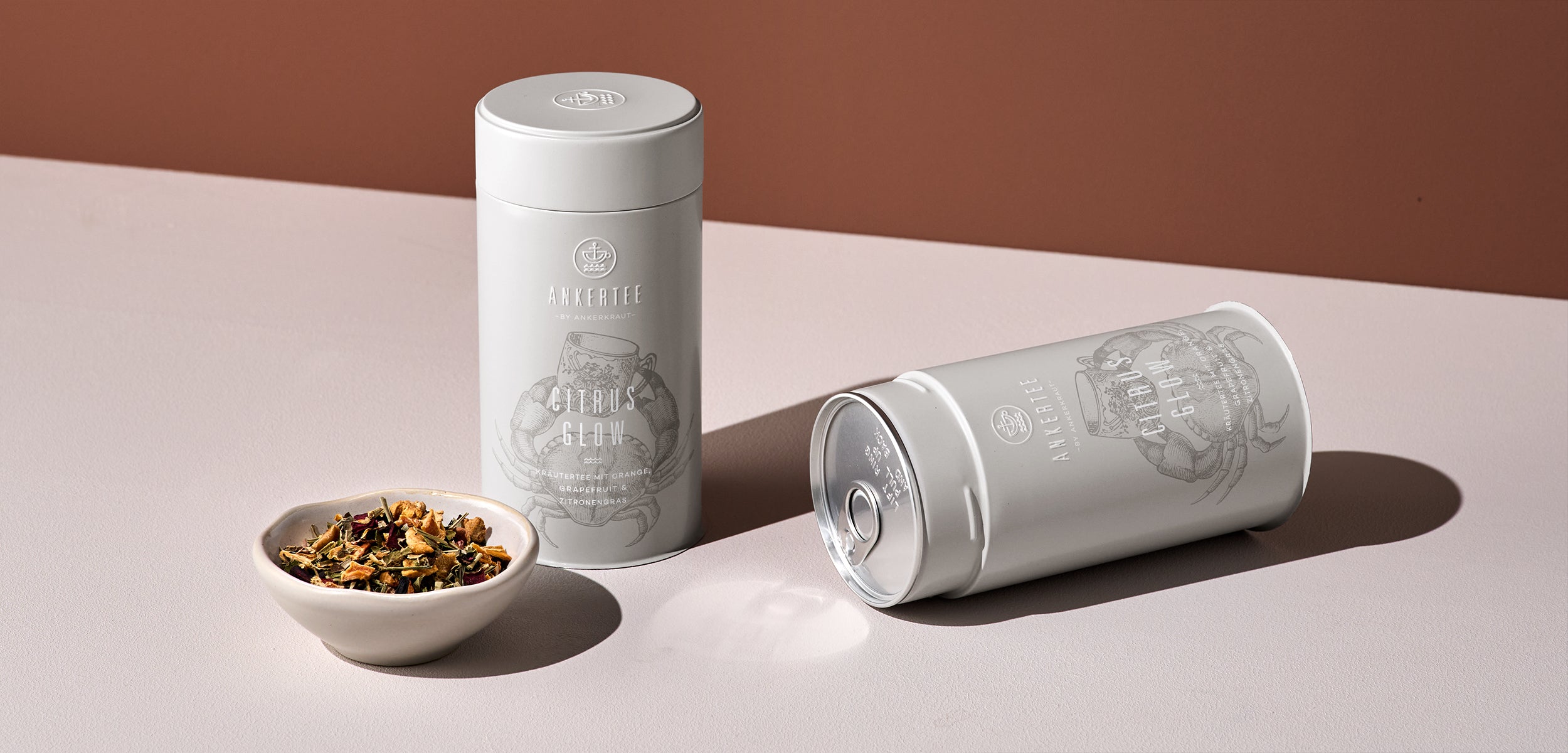 Eine stehende und eine liegende Teedose von Ankertee mit der Aufschrift CITRUS GLOW, davor eine Schale mit loser Teemischung.