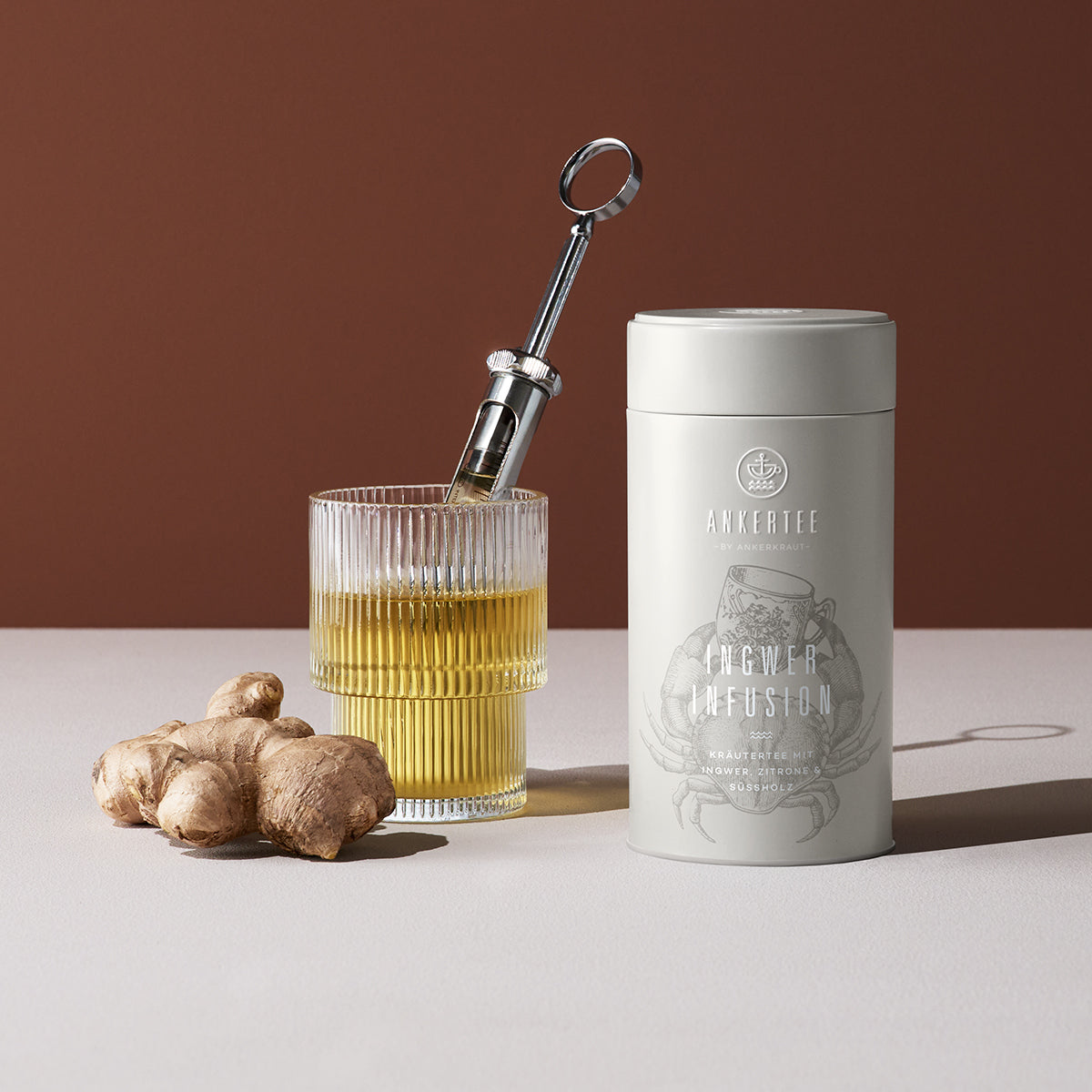 Eine Ankertee-Dose mit der Aufschrift INGWER INFUSION, daneben ein gefülltes Teeglas mit einer Infusionsspritze und davor eine Ingwerknolle