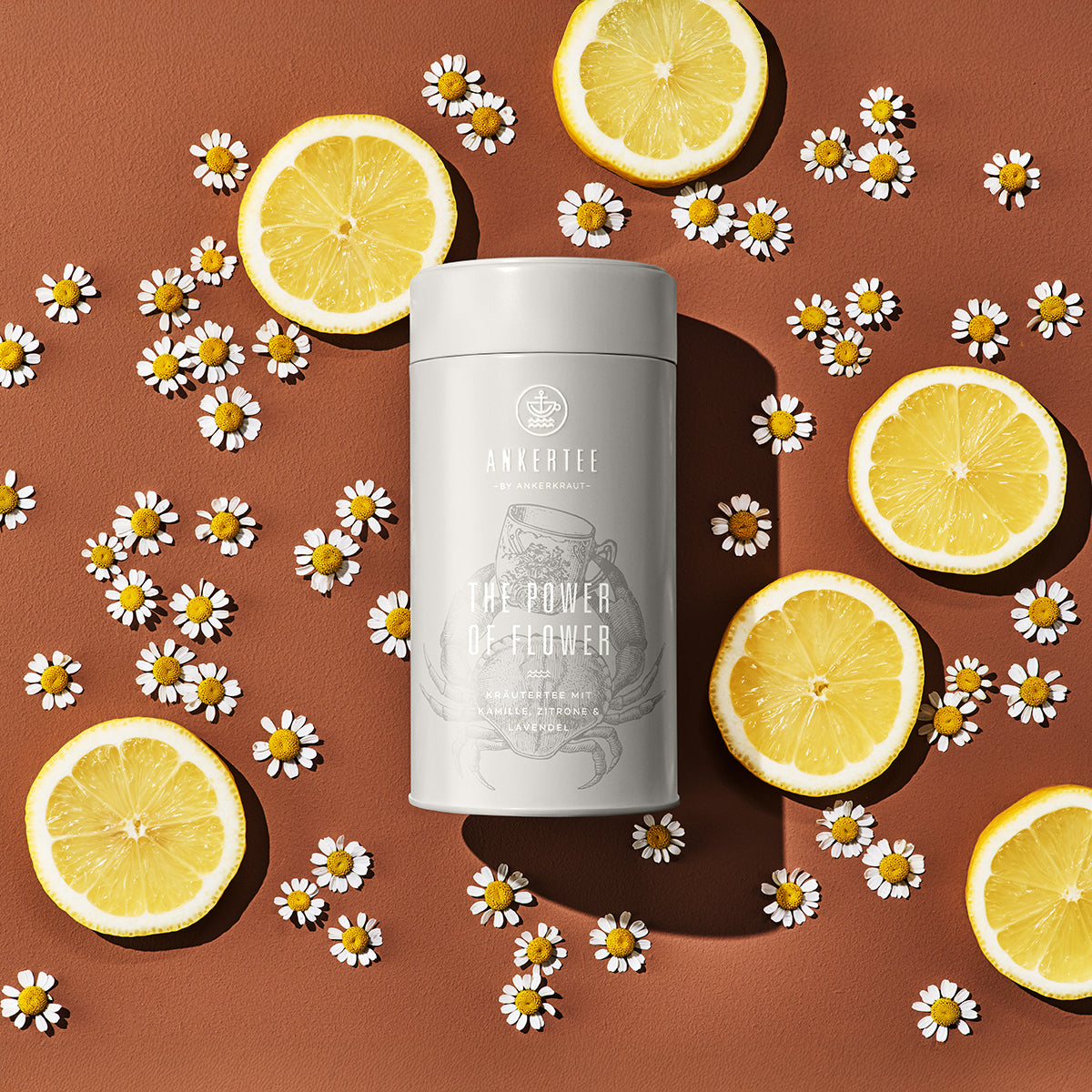 Eine Ankertee-Dose mit der Aufschrift THE POWER OF FLOWER liegt auf einer braunen Fläche umgeben von Blüten und Zitronenscheiben